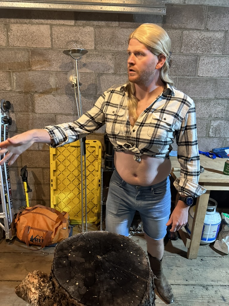 Guest in a blond mullet wig, knotted flannel crop top, jorts, and cowboy boots in the garage