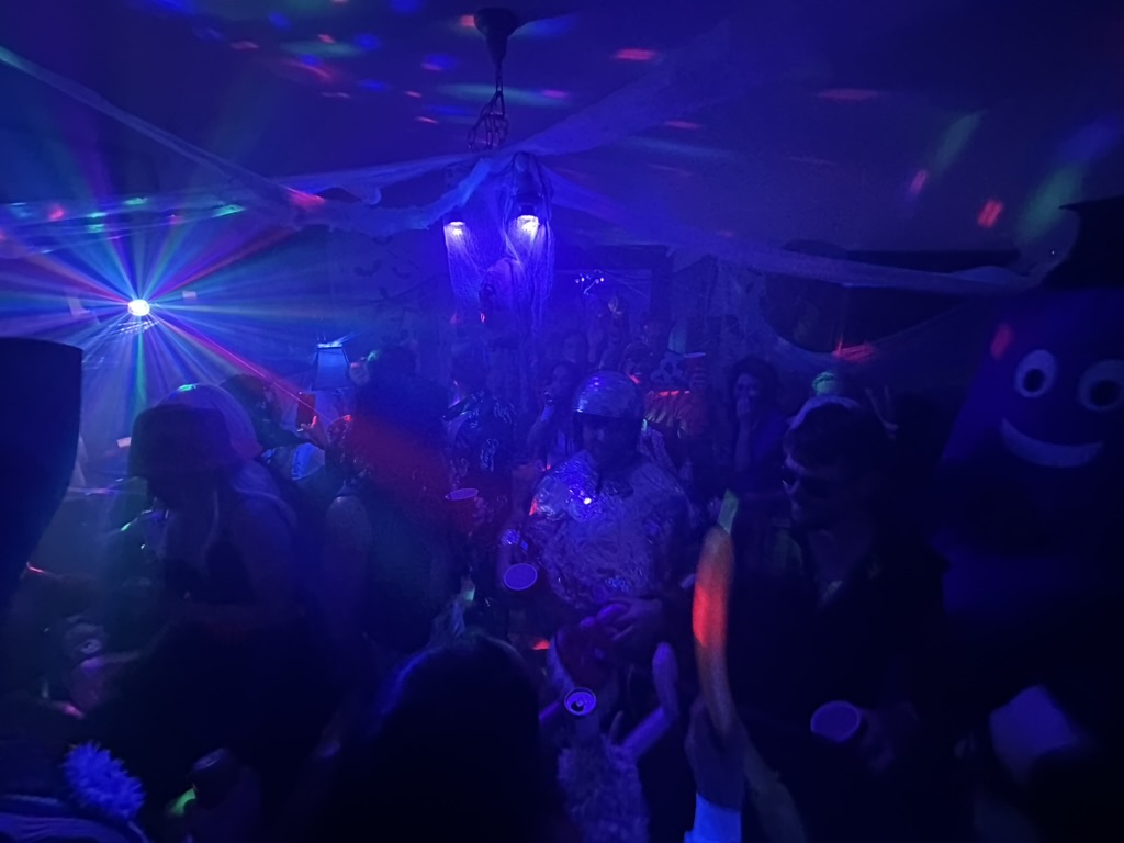 Packed dance floor with disco lights, cobwebs, and a purple mascot costume in the crowd