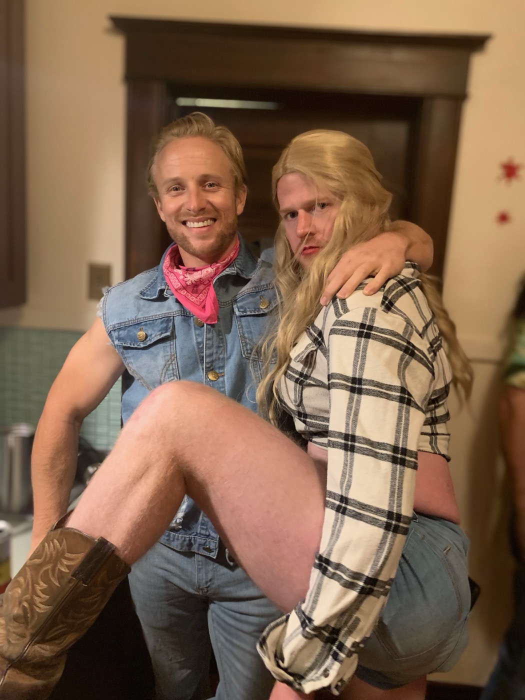 A guest in a denim vest and cowboy boots poses with the blond mullet guest draped over his knee