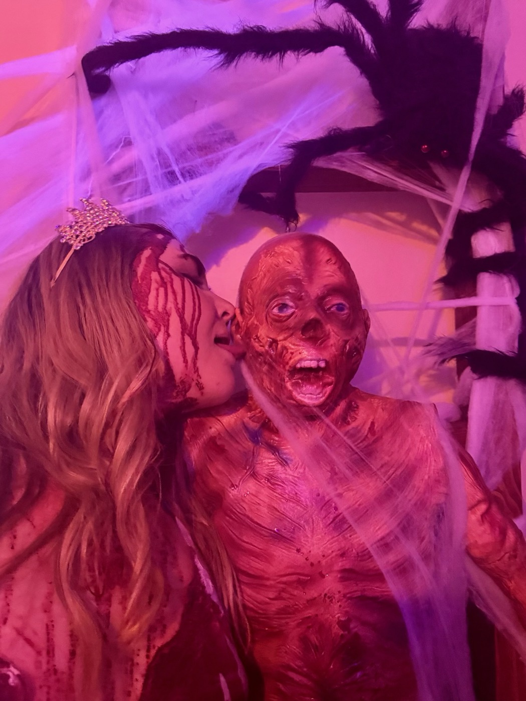 A guest in a tiara leans in to kiss a gruesome zombie-monster mask beneath a giant spider