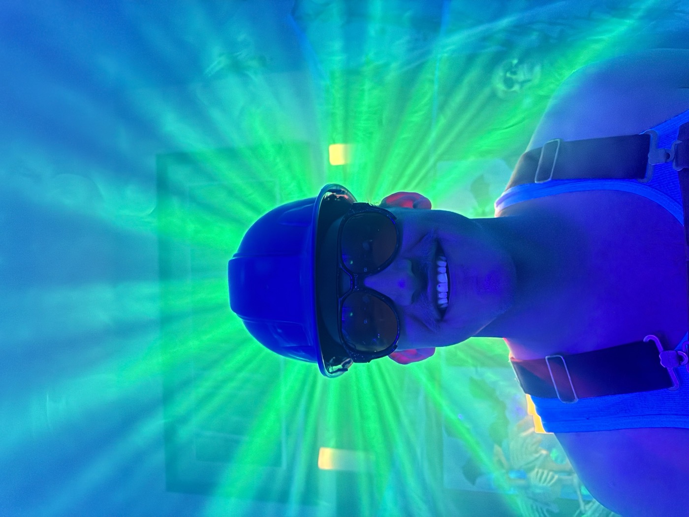 A grinning guest in a hard hat and sunglasses lit by green laser beams